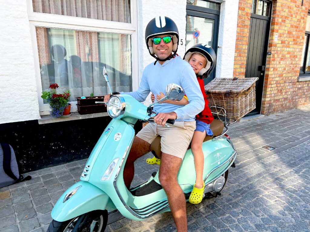 A man and a child, both wearing helmets, sit on a light blue scooter parked on a cobblestone street in front of a white and brick building. The child is sitting behind the man and smiling at the camera.