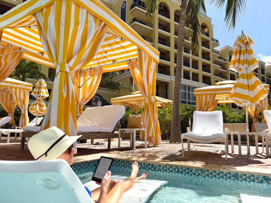 A person in a sunhat relaxes on a lounge chair in a pool, reading an e-reader. Yellow and white striped cabanas and lounge chairs surround the area, with a resort building and palm trees in the background.