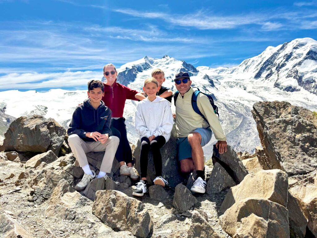 A family of five poses on rocky terrain with snow-covered mountains and a bright blue sky in the background. All are dressed in casual, sporty clothing, smiling and sitting close together.
