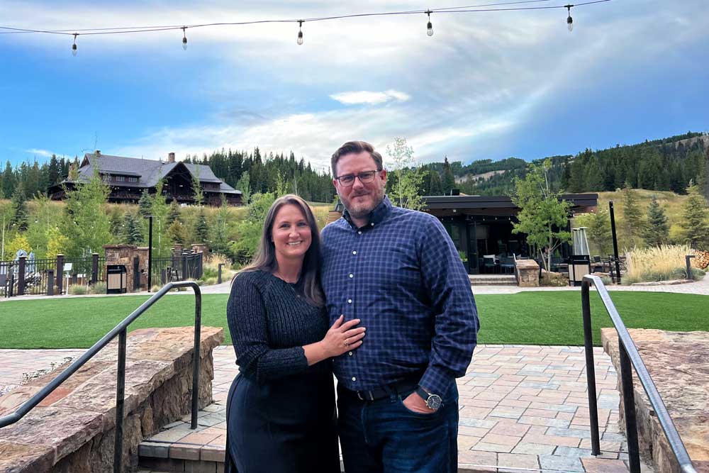 A smiling couple stands together outdoors on a paved patio with green grass, surrounded by trees, a rustic lodge, and string lights overhead under a partly cloudy sky.