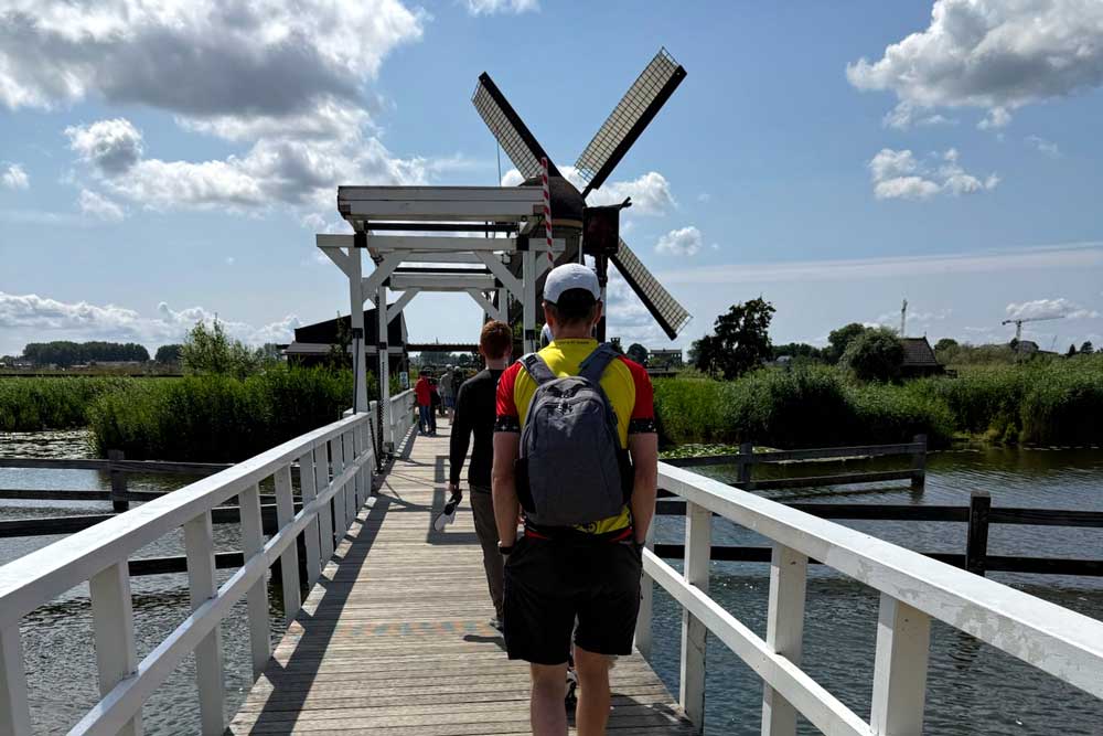 People walk on a wooden bridge over water toward a traditional windmill, surrounded by greenery and under a partly cloudy sky. The person in front wears a backpack and sporty clothes.