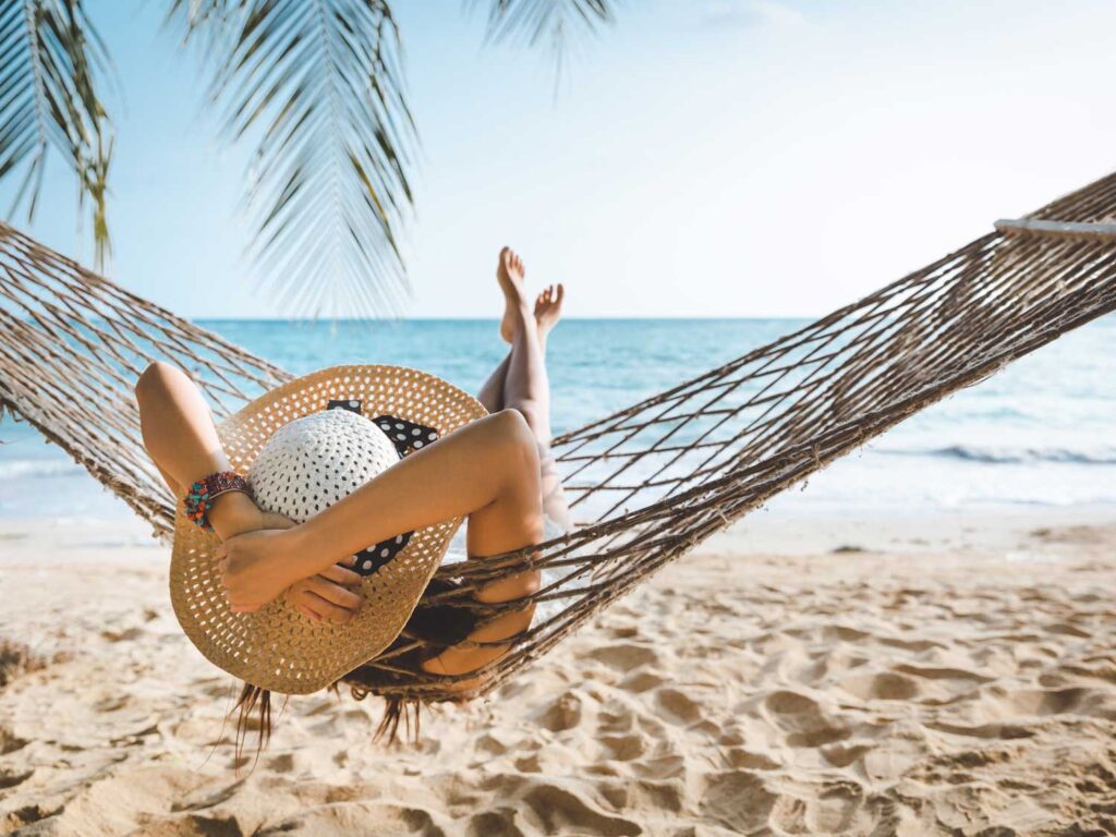 A person relaxing on a hammock at the beach, wearing a wide-brimmed hat and holding it with both hands, with palm leaves overhead and the ocean in the background.